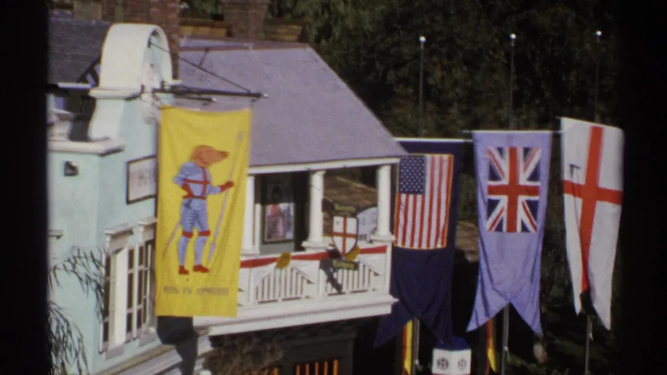 1972 Arizona frame showing a miniature village attraction facade with a yellow cartoon banner, U.S. flag, Union Jack, and red cross flag.