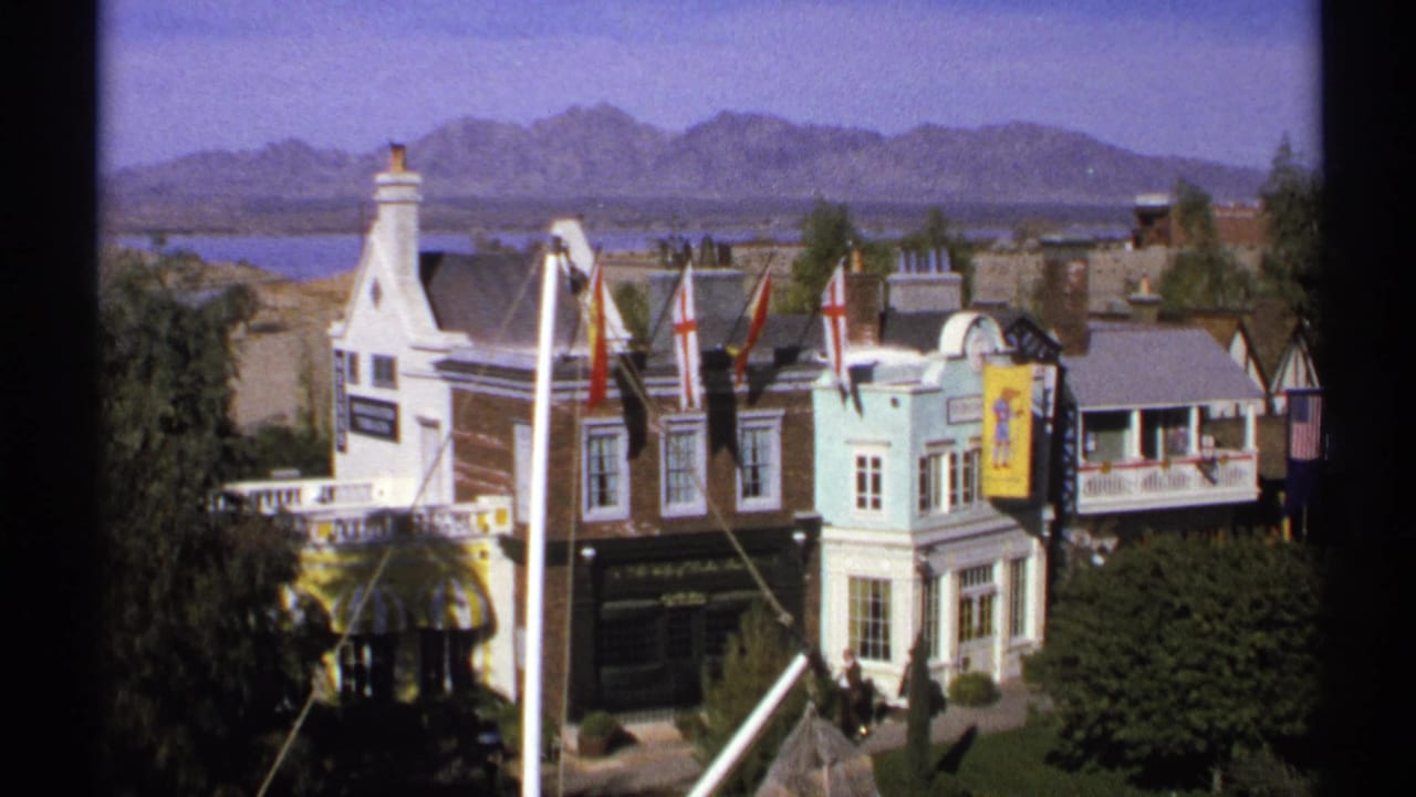1972 Arizona 8mm frame showing colorful miniature town facades, flags, and distant mountains beyond water.