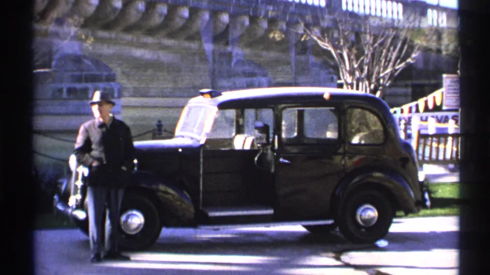 1972 Arizona home-movie frame of a man in a fedora near a classic black sedan, with a stone bridge and railings behind him.