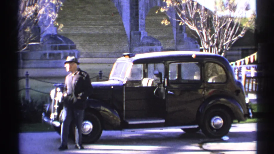 1972 Arizona frame showing a fedora-wearing man beside a classic sedan under stone bridge arches, with railings and a bare tree in sunlight.