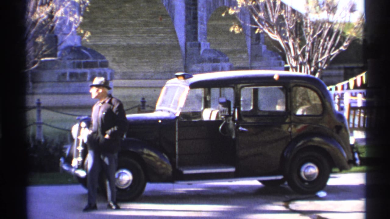 Man in a fedora stands beside a black vintage car with its door open near stone bridge arches.