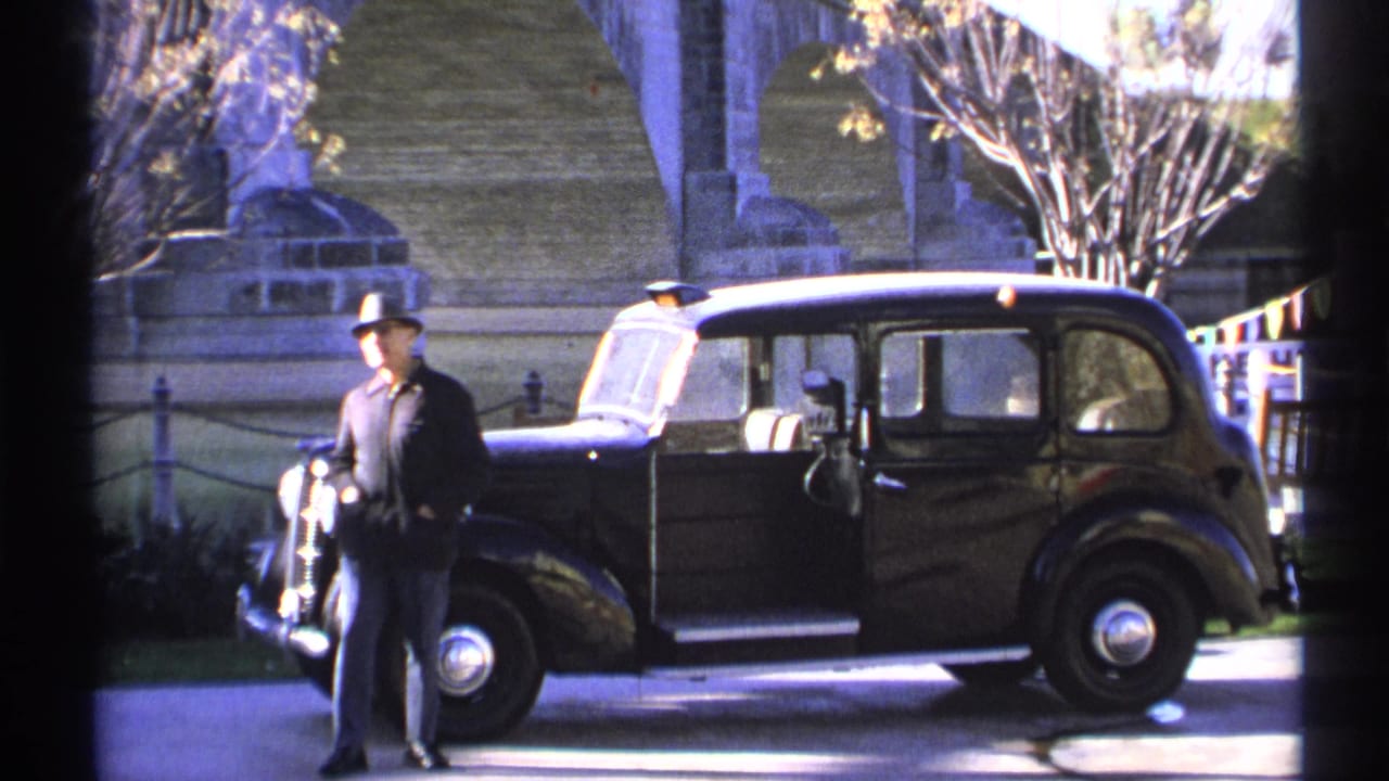 Man stands in front of a black vintage car with an open door and stonework in the background.