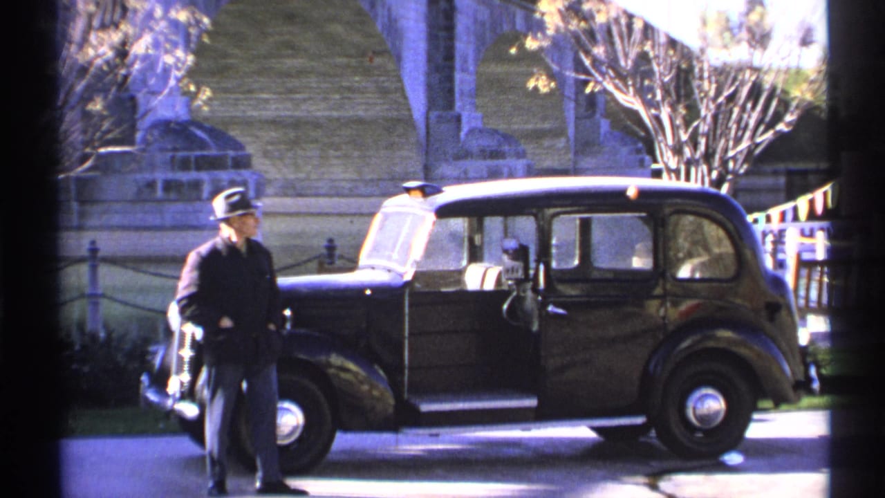 Man in a fedora stands by the front of a black vintage car with an open door.