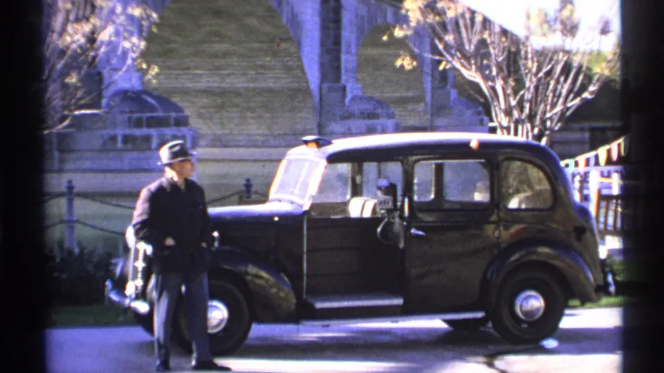 1972 Arizona 8mm frame showing the man beside the front of the black car, with the open door, chrome details, bridge stonework, and trees visible.