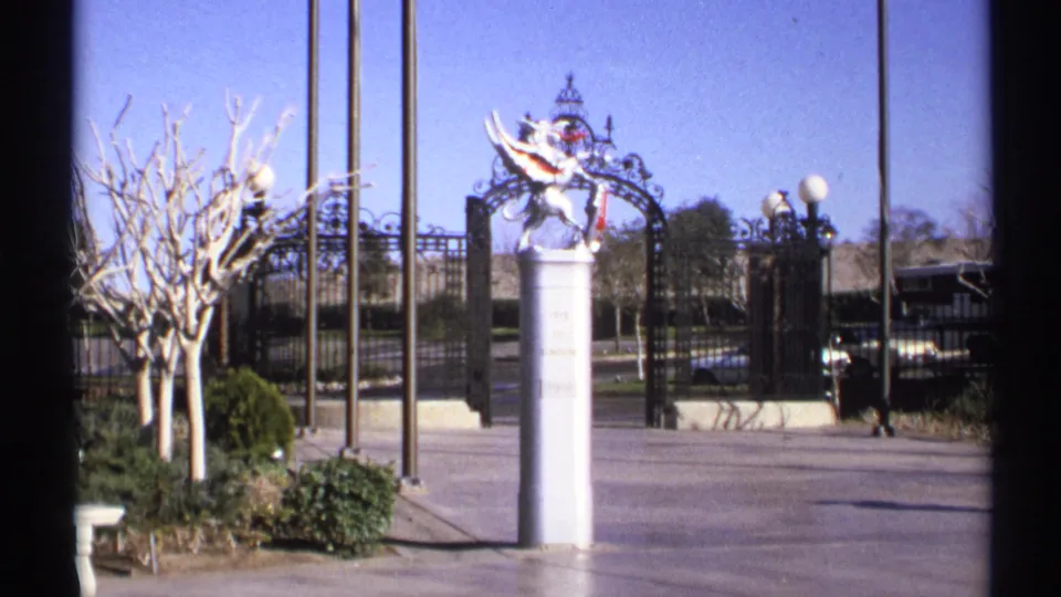 1972 Arizona 8mm frame showing a decorative iron gate, flagpoles, formal landscaping, bare trees, and part of a dark car at the right edge.