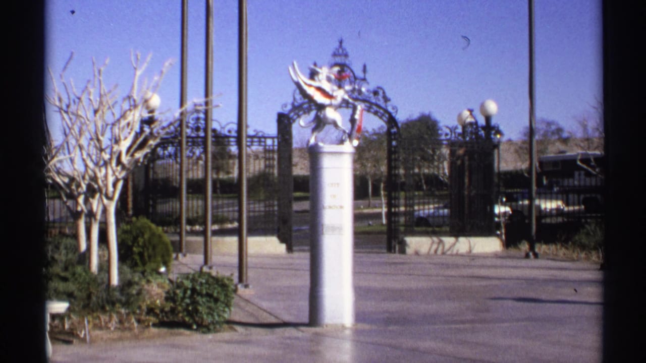 White pedestal statue beside an ornate gate with bare trees and globe lamps.