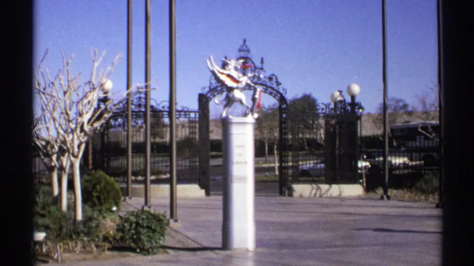 1972 Arizona home-movie frame with a white winged statue on a pedestal, black iron gate, lamps, shrubs, and bare trees.