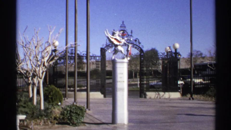 1972 Arizona 8mm frame showing the statue and gate from a nearby angle, with winter-bare trees and lamp posts framing the entrance.