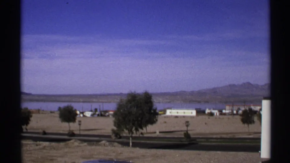 1972 Arizona home-movie frame opening to a wide desert landscape with calm water, distant mountains, scattered buildings, and trees.