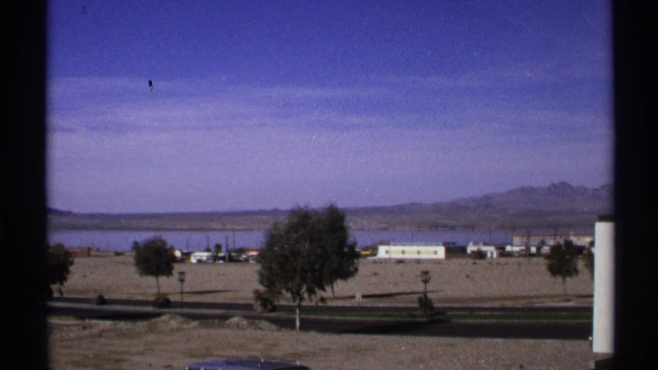 Black car partly visible on a road below desert lots, low buildings, trees, water, and distant mountains.