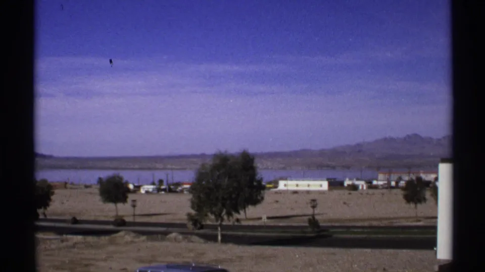 1972 Arizona 8mm frame showing a sparse roadside area with trees, low buildings, water, and mountains beyond a road where a dark car is partly visible.