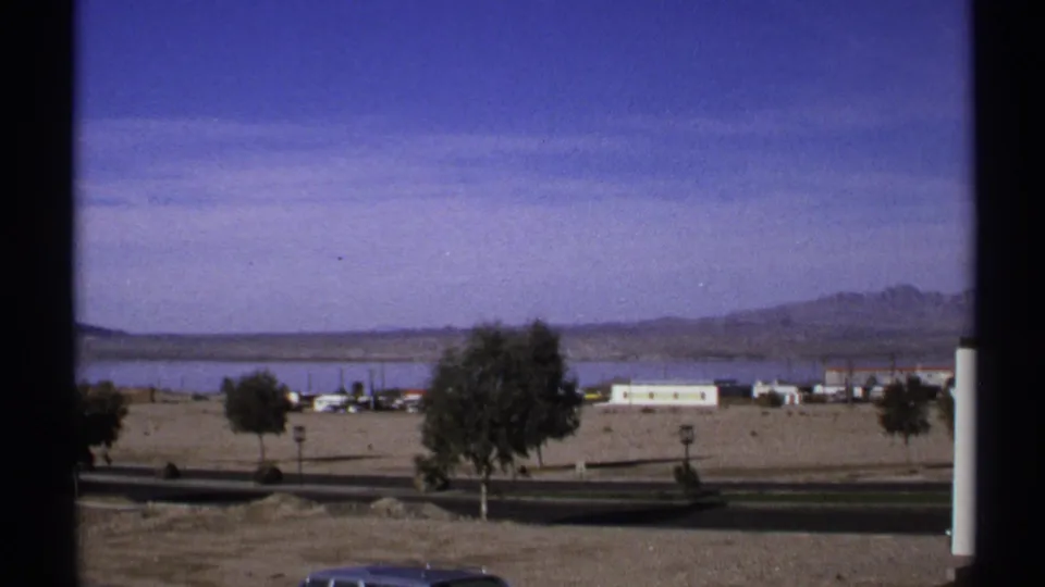 1972 Arizona home-movie frame of a near-empty road below low roadside buildings, desert ground, scattered trees, water, and distant mountains.