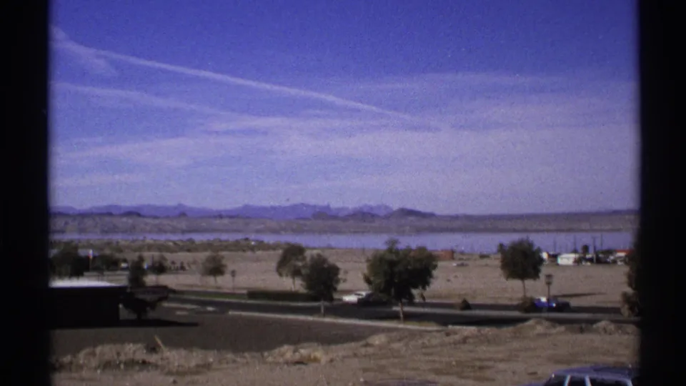1972 Arizona 8mm frame with a wider roadside view, showing cars along the lower road, open desert ground, low buildings, water, and mountains.
