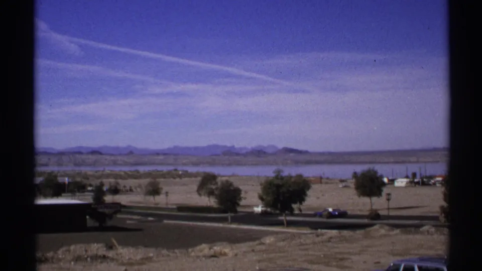 1972 Arizona frame showing a dark car traveling along a road beneath an open desert view with trees, low buildings, water, and distant mountains.