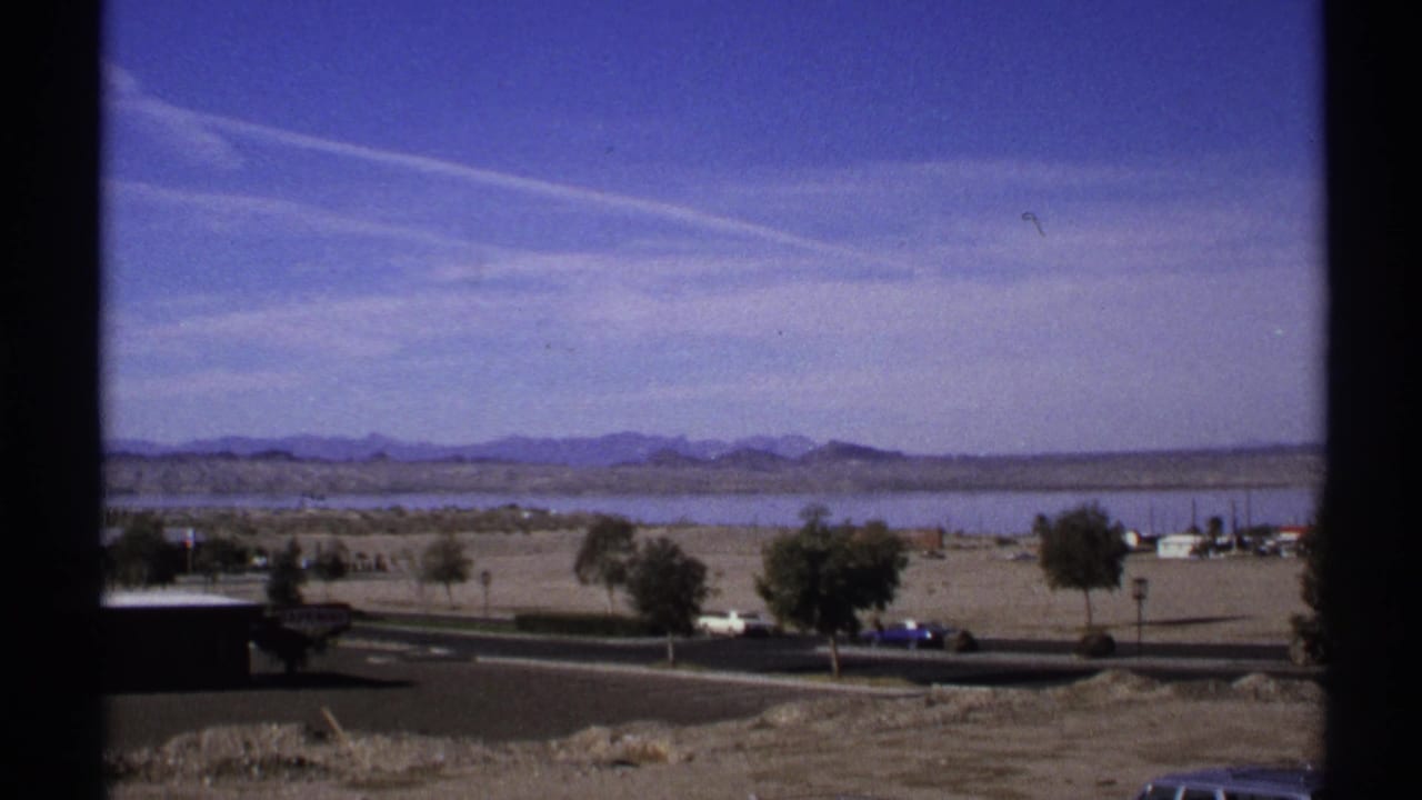 Desert lake with road, trees, vehicles, and distant mountains under a blue sky.