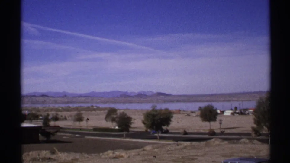 1972 Arizona home-movie frame looking across a roadside foreground to a broad lake, sparse trees, small vehicles, and a mountain horizon.