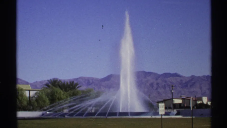 1972 Arizona 8mm frame of a tall fountain plume rising from a roadside water feature, with palm trees, signs, low buildings, and mountains behind it.