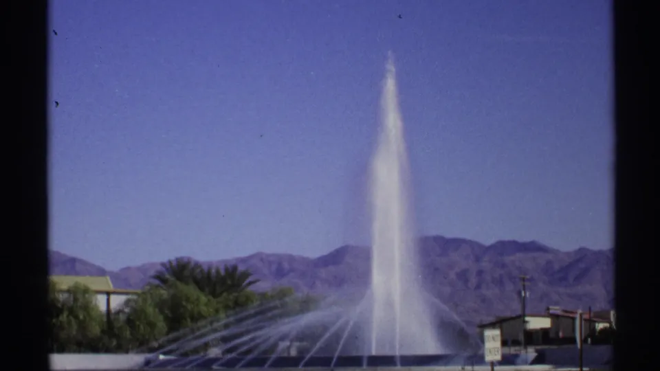 1972 Arizona home-movie frame showing a fountain shooting water upward beside palms and roadside structures, with mountains visible beyond.