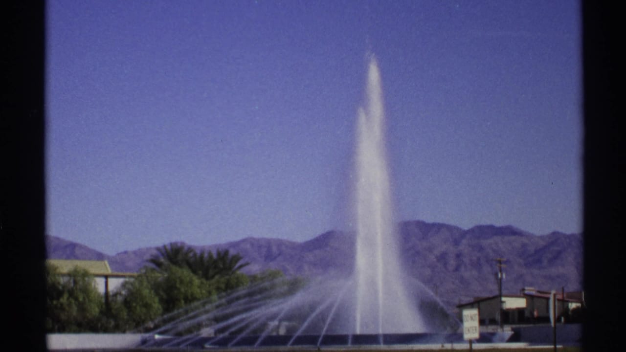 Tall water fountain rising in front of desert mountains and palm trees.
