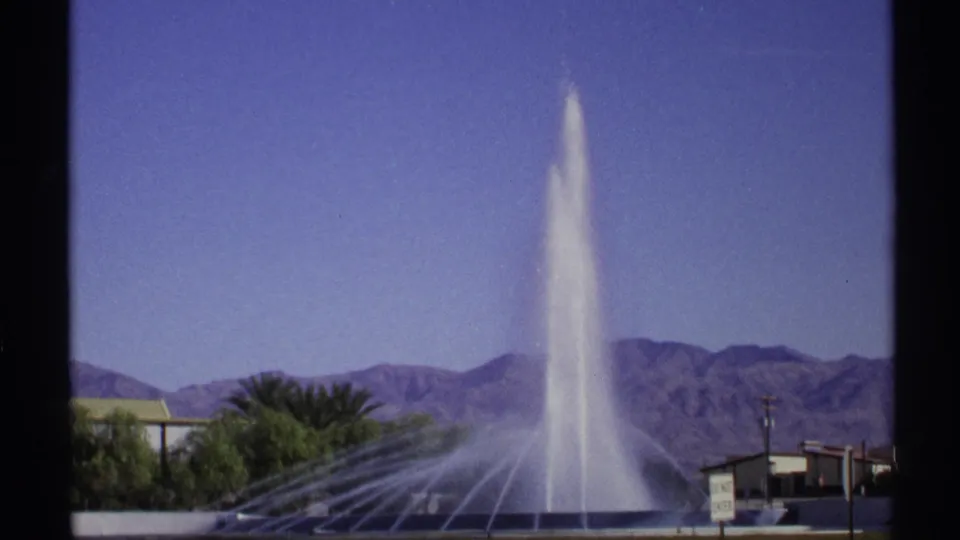 1972 Arizona 8mm frame showing a tall fountain against blue sky, desert mountains, palms, and low roadside buildings.