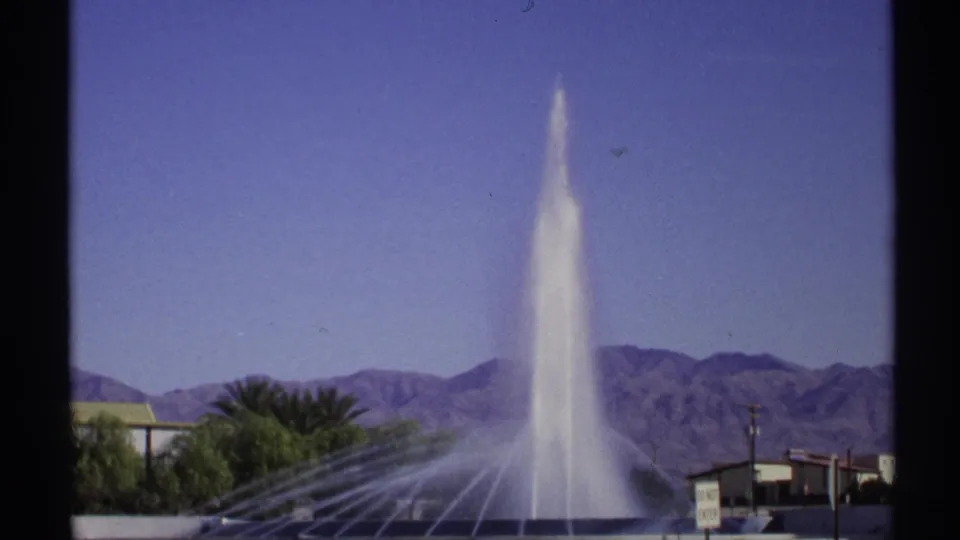 1972 Arizona home-movie frame of a fountain plume and side sprays set before rugged desert mountains and low buildings.