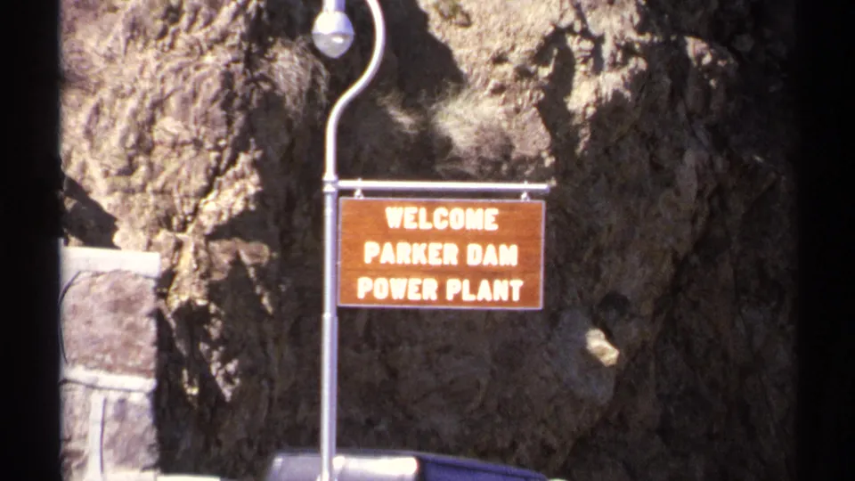 1972 Arizona frame showing a lamp post and wooden Welcome Parker Dam Power Plant sign against a rocky cliff face.
