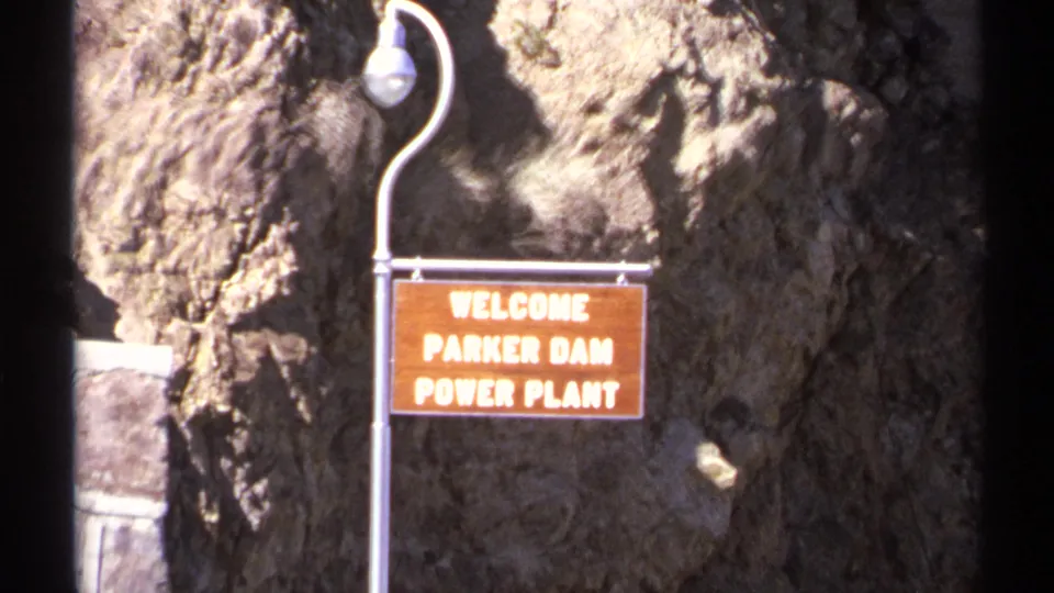 1972 Arizona 8mm frame with a readable Parker Dam Power Plant welcome sign, curved lamp post, and rough rock wall behind it.