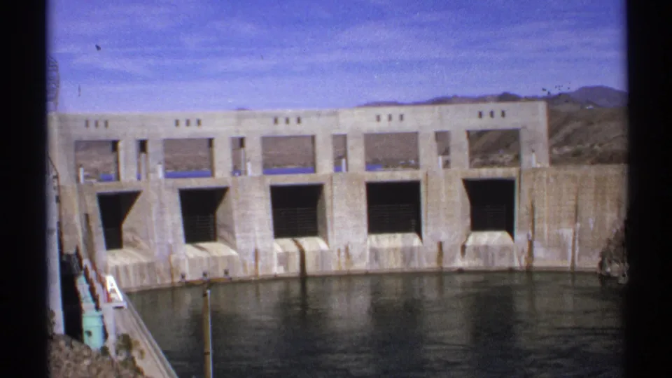 Wide 1972 Arizona 8mm view of the Parker Dam Power Plant facade, water below, and low desert hills beyond.