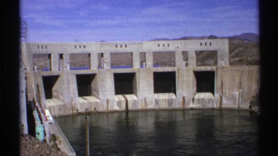 1972 Arizona home-movie frame with multiple dam gate openings, a concrete wall, reservoir water, and desert hills.