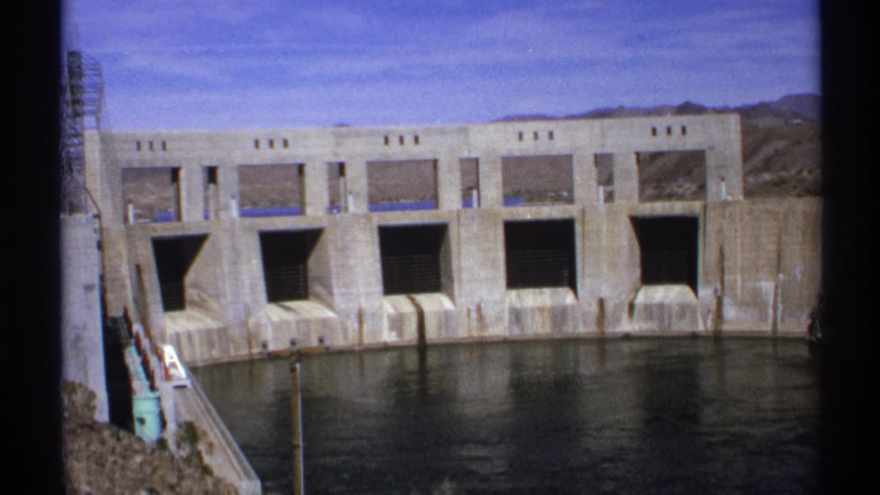 Concrete dam face with dark spillway openings above calm water in a desert setting.
