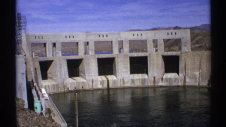 1972 Arizona 8mm frame showing a massive concrete dam, spillway openings, calm water, and arid hills beyond.