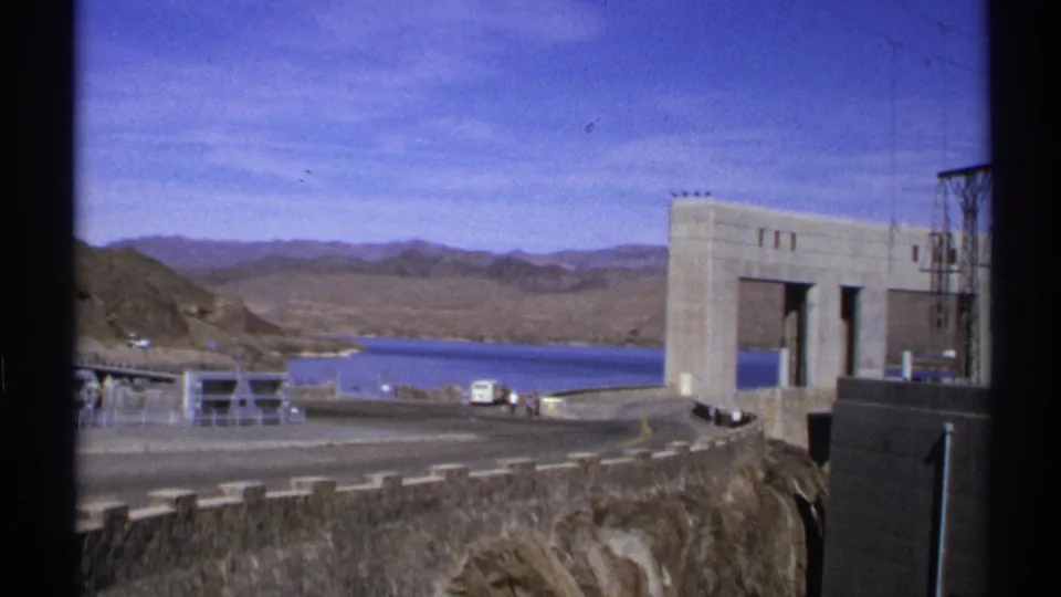 1972 Arizona frame after the pan, showing reservoir water, desert mountains, roadway, vehicles, tiny visitors, and part of the dam structure.