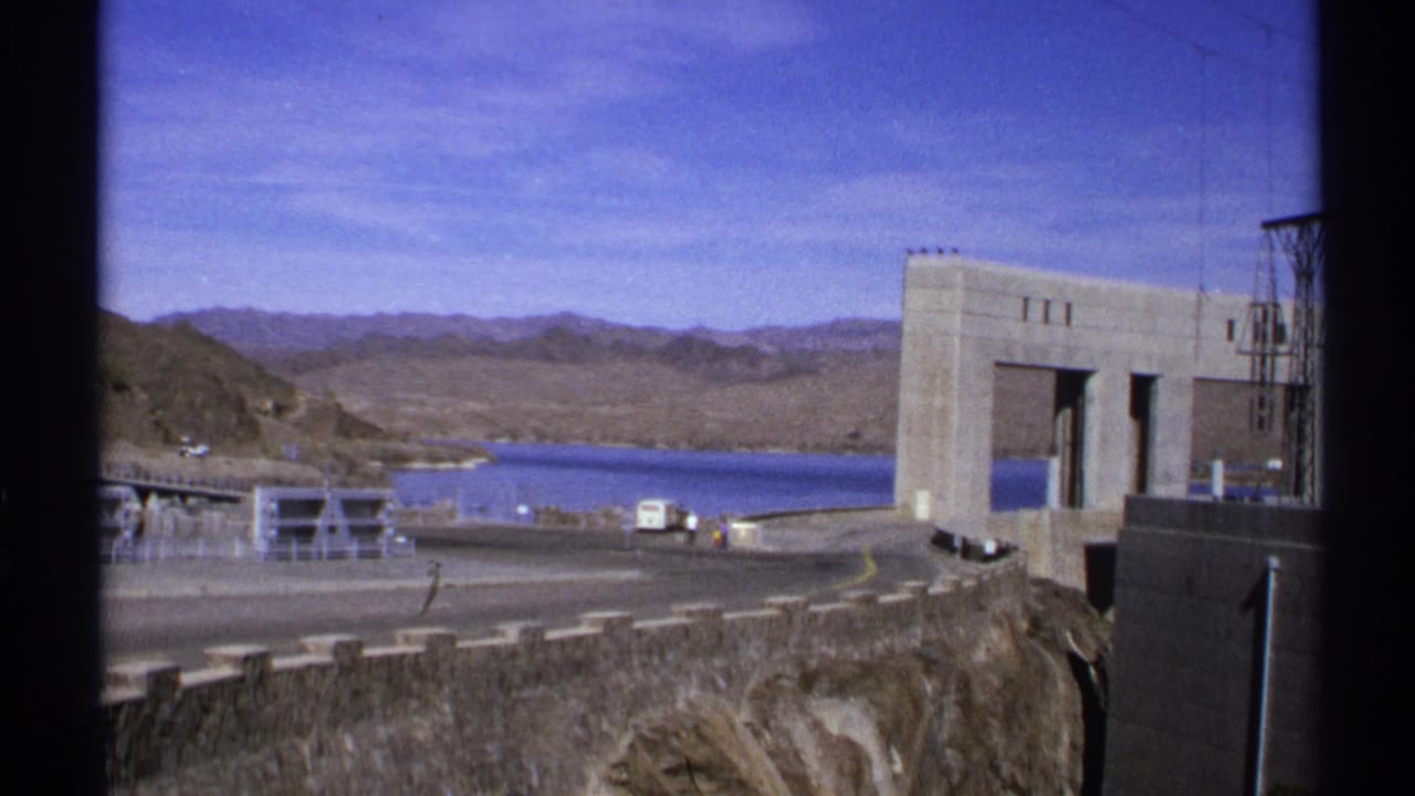 1972 Arizona 8mm frame showing a concrete dam, blue reservoir water, desert hills, road barriers, and small vehicles near the overlook.