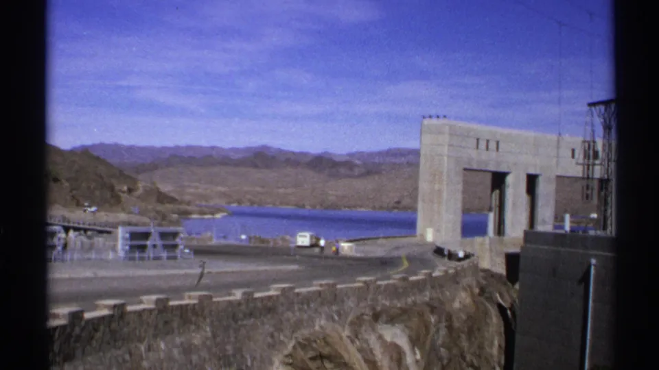 1972 Arizona 8mm frame showing a concrete dam, blue reservoir water, desert hills, road barriers, and small vehicles near the overlook.