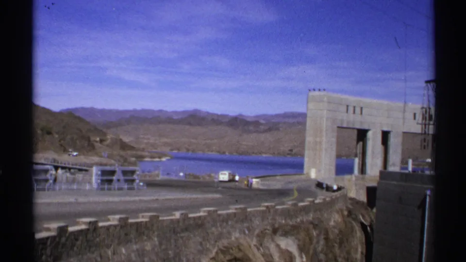 1972 Arizona home-movie frame with the dam structure at right, reservoir and mountains beyond, and a road with distant vehicles below.