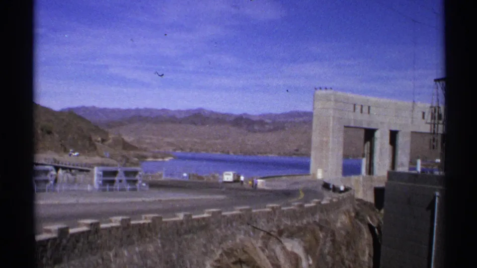1972 Arizona 8mm frame showing the concrete dam at right, a blue reservoir, desert mountains, roadway barriers, and rocky terrain in the foreground.