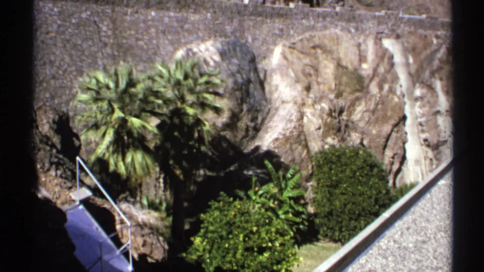 1972 Arizona visitor-area frame looking down at palms, shrubs, rock formations, steps, metal railing, and a surrounding wall near the dam site.