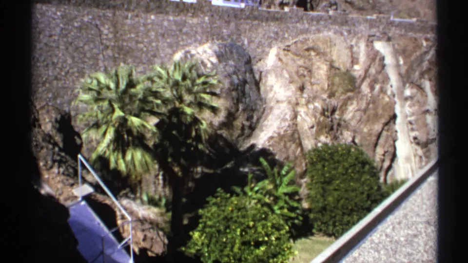 1972 Arizona 8mm frame showing palms, shrubs, rock face, stone retaining wall, and a railed walkway beside a dam.