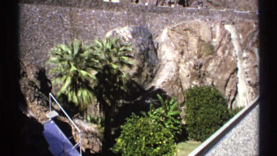 1972 Arizona 8mm view of palms and shrubs below a rugged rock face, with a railed sloped walkway at the dam.