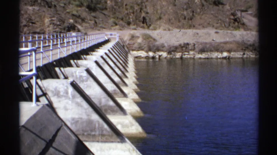 1972 Arizona frame showing the dam's concrete side, metal guardrails, rocky bank, and calm blue reservoir water.