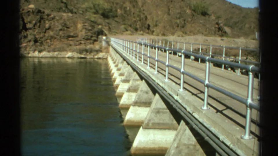 1972 Arizona 8mm frame looking along a concrete dam or bridge walkway with metal railings over calm green-blue river water.