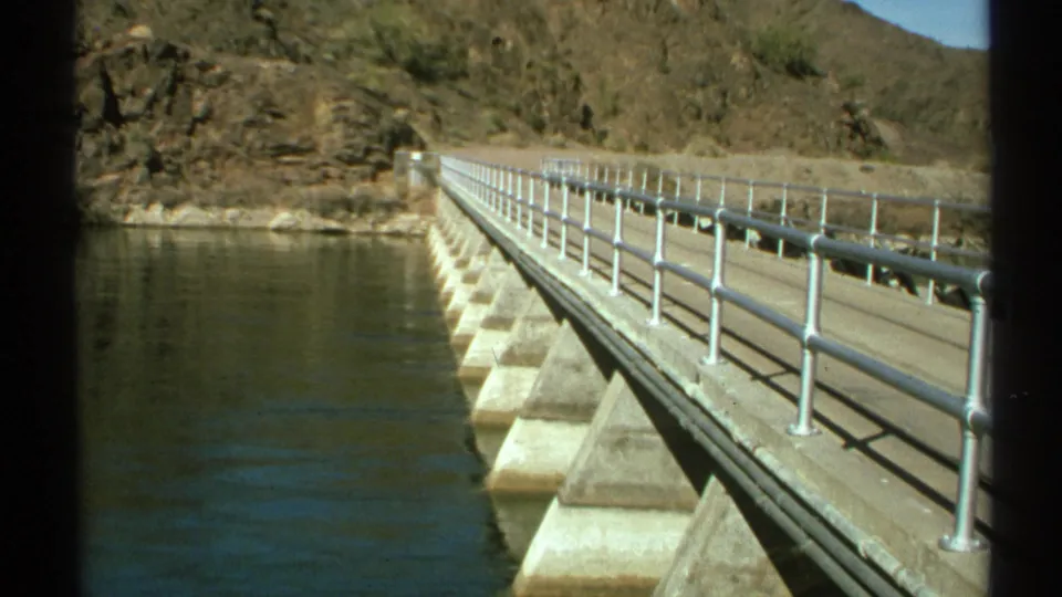 1972 Arizona 8mm view along a concrete bridge or dam walkway, showing metal railings, repeating supports, river water, and desert rock slopes.
