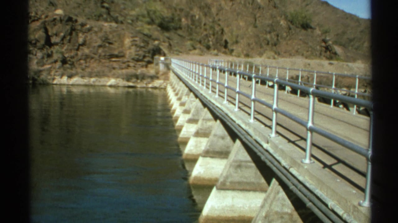1972 Arizona 8mm frame showing a concrete dam or bridge span over water, with metal railings and rocky desert terrain beyond.