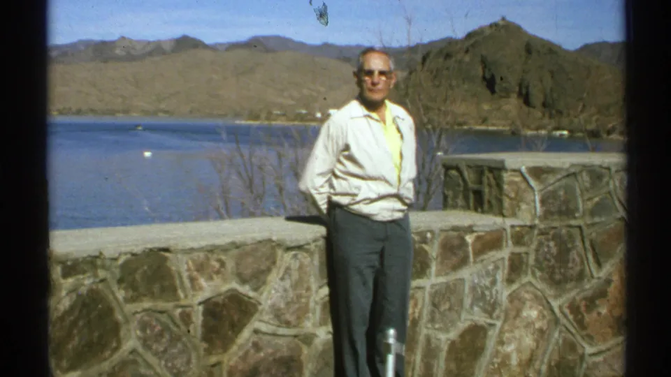 1972 Arizona home-movie frame of an adult man posing at a stone overlook, with a blue lake and rugged desert mountains in the background.