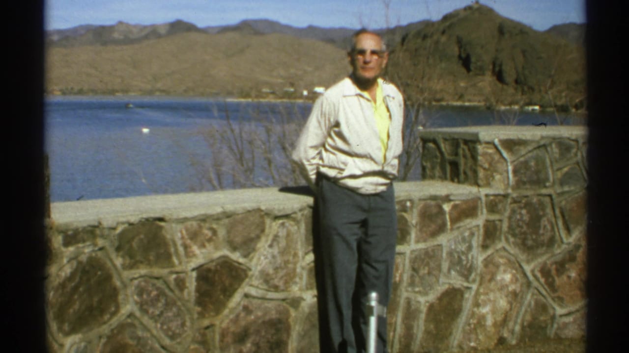 Older man in sunglasses stands by a stone wall overlooking a lake and desert mountains.