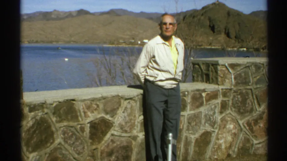 In 1972 Arizona home-movie footage, an older man poses in sunglasses beside a stone lakeside overlook with arid mountains beyond.
