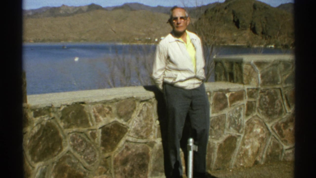 Older man stands with hands behind his back near a stone wall, lake, and mountains.