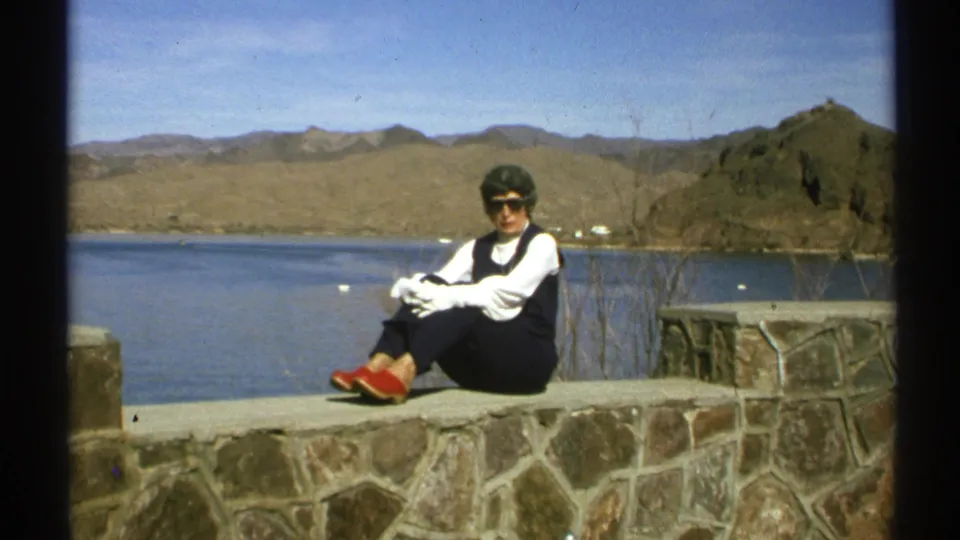 In 1972 Arizona footage, an older woman wearing sunglasses, a dark vest, and red shoes sits on a stone wall by a desert lake.