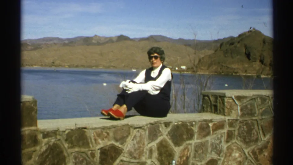 An older woman sits for the camera at an Arizona lakeside overlook, framed by stonework, blue water, and dry mountain ridges.