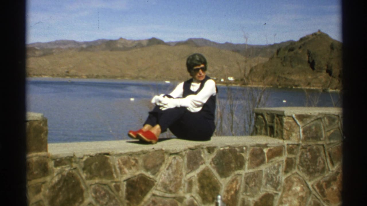 Woman in sunglasses sitting on a stone wall beside a lake with desert hills behind her.